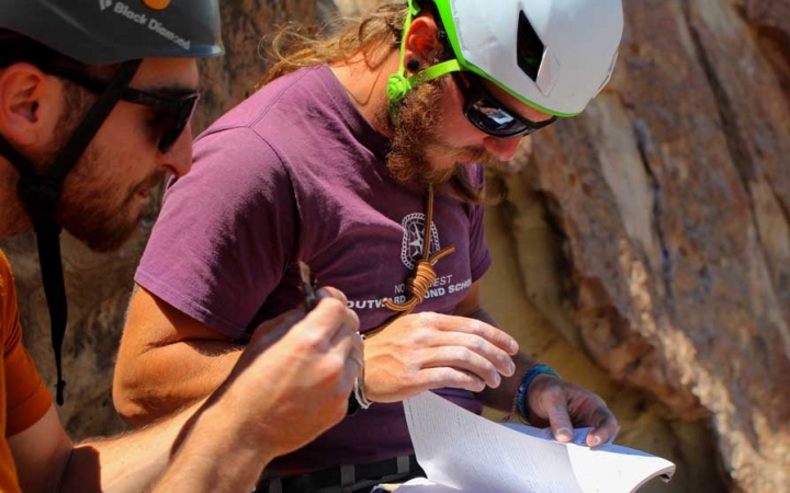 two people wearing helmets examine a book whiles standing next to a rock wall
